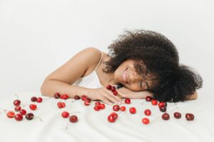 Happy woman with natural curls smiling among scattered cherries on a white background.