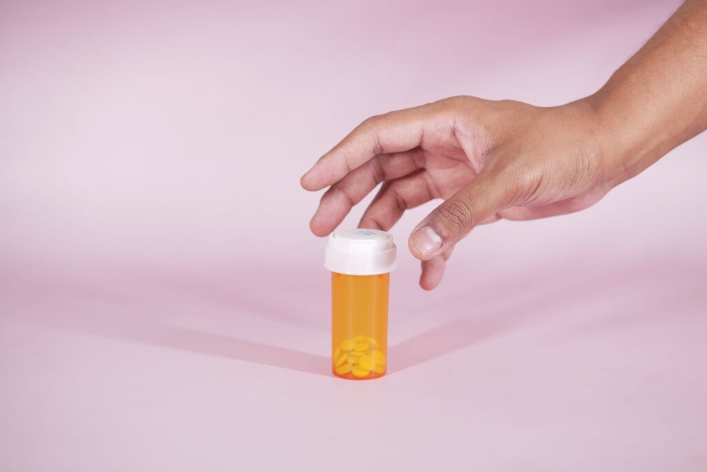 A hand reaches for a prescription bottle with yellow pills on a pink background, symbolizing healthcare.