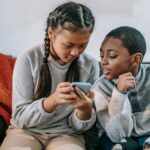 Attentive Asian girl with black boy sharing cellphone while resting on couch in house