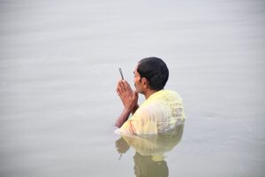 A man standing in waist-deep water with hands in prayer at dawn, symbolizing spiritual devotion.