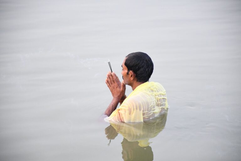 A man standing in waist-deep water with hands in prayer at dawn, symbolizing spiritual devotion.