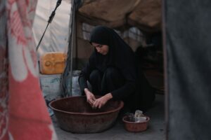 A Middle Eastern woman in hijab washes dishes in a makeshift Gaza tent.