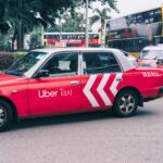 Red Uber taxi parked on a bustling city street with buses and greenery in the background.