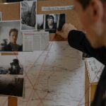 Detective examining a crime investigation board with photos, maps, and notes indoors.