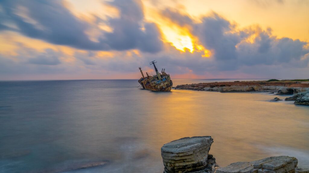 A serene view of a shipwreck near a rocky shore at sunset with dramatic sky.