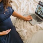 From above of crop anonymous expectant female caressing belly while surfing internet on netbook on cozy bed