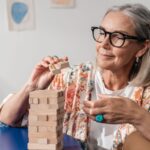 Elderly woman having fun with a Jenga puzzle indoors, wearing stylish eyewear.