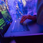 Close-up of hands typing on a laptop displaying cybersecurity graphics, illuminated by purple light.