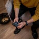 Man in yellow sweater using remote to control robotic vacuum on wooden floor.