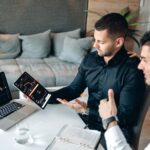 Two businessmen discuss digital trading insights with laptop and tablet in a modern office setting.