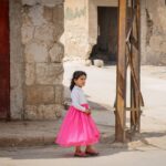 A young girl stands in a Syrian street, wearing a bright pink dress. Captured in Idlib.