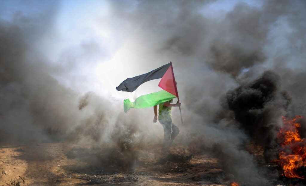 A protestor carrying a Palestinian flag amidst smoke and flames in Gaza.