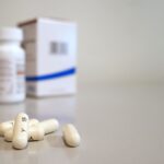 Close-up image of white capsules with medicine bottle and box in the background.