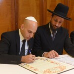 Jewish men in suits sitting at a desk, reviewing and signing a document.