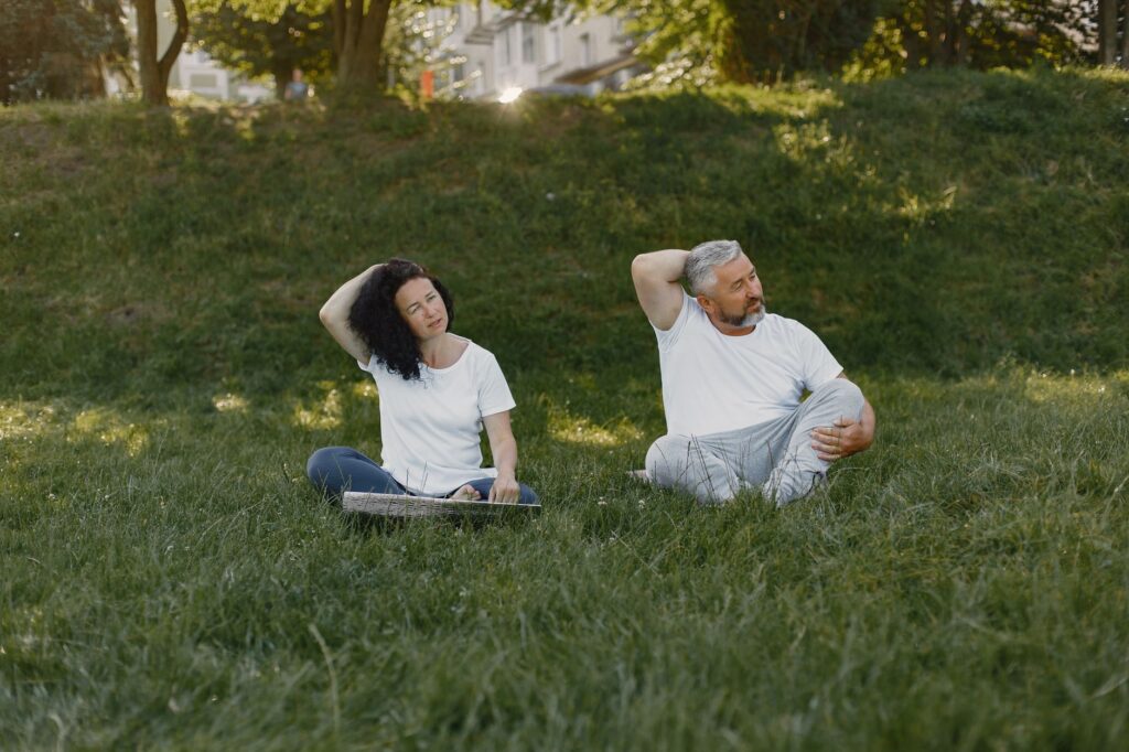 Couple doing neck stretches outdoors, promoting a healthy lifestyle in a sunny park setting.