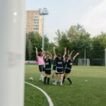 A women's soccer team in black uniforms celebrates a victory on an outdoor soccer field.