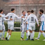 A group of soccer players in white uniforms celebrating on a soccer field during a daytime match.