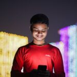 Smiling young man in red shirt using smartphone against a backdrop of festive lights at night.
