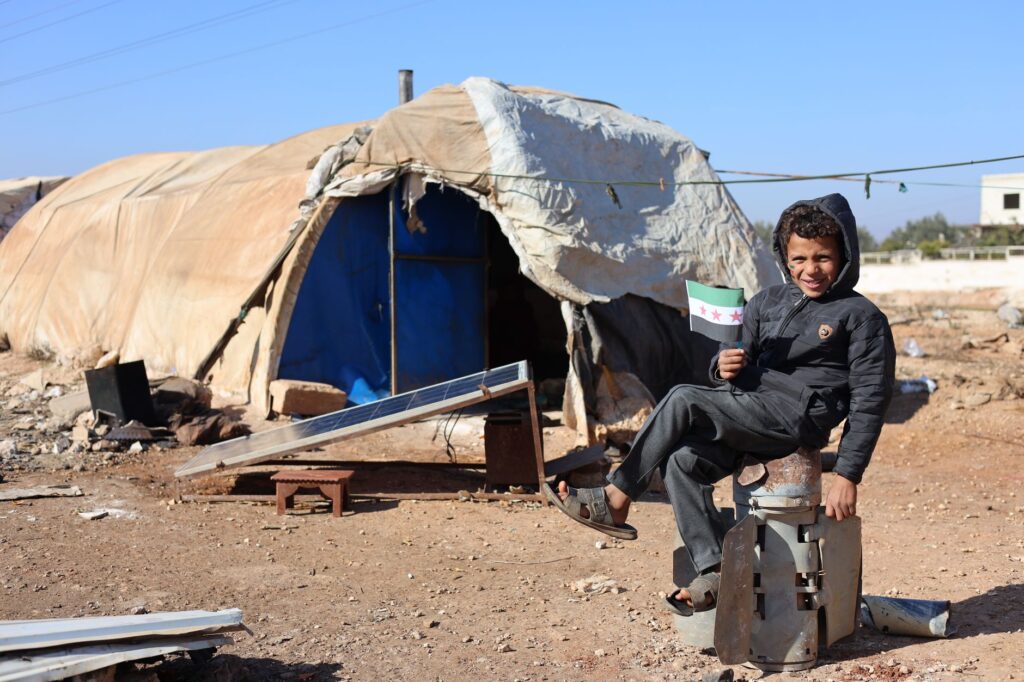 Young boy sits cheerfully outside a tent in Idlib holding a flag, symbols of hope.