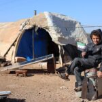 Young boy sits cheerfully outside a tent in Idlib holding a flag, symbols of hope.