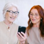 Happy senior women looking at screen of mobile phone while watching video through internet