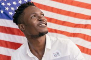 Man smiling with a voting sticker against an American flag background, symbolizing patriotism.
